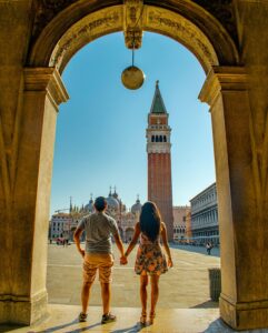 Pareja viajera disfrutando de la vista frente a una plaza en Venecia, con un minarete histórico como fondo, destacando la mezcla de culturas y arquitectura en la ciudad