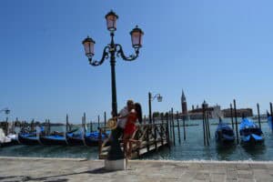 Pareja viajera caminando junto a un canal en Venecia con góndolas al fondo – Viajeros por el Mundo